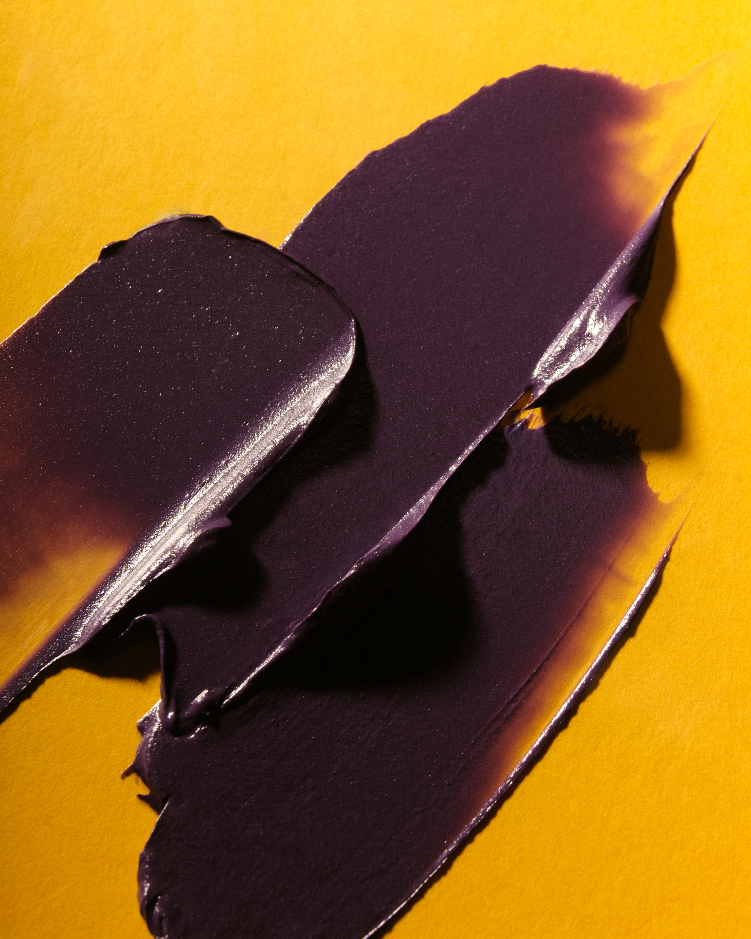 Traces épaisses de texture cosmétique violette, appliquées en couches superposées sur un fond jaune.
