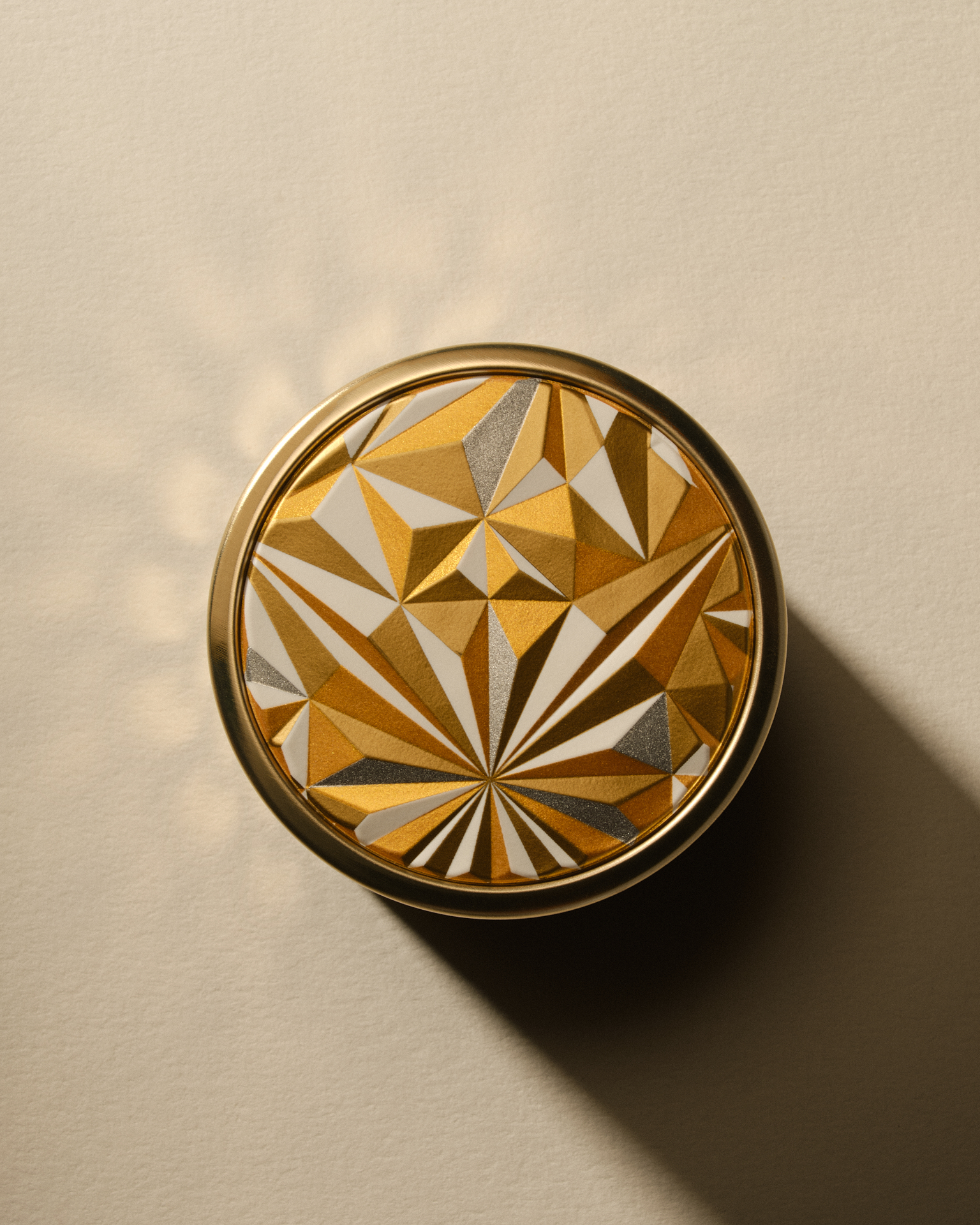 boîtier cosmétique doré, vu de dessus, présentant un motif géométrique en relief, éclairé par une lumière directionnelle.